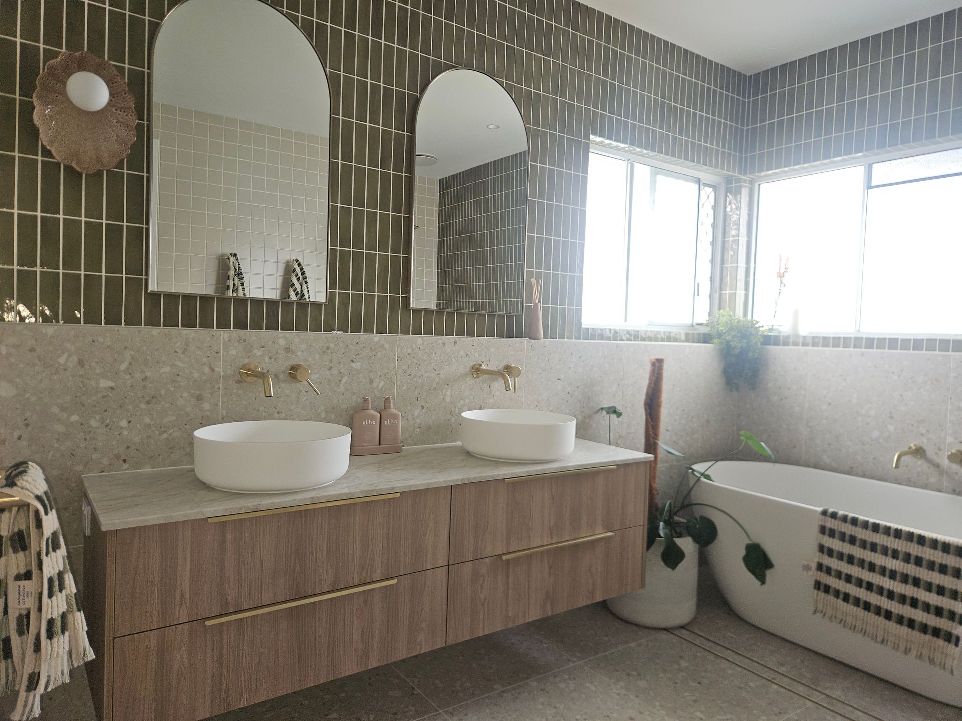 Bathroom with wood vanity, round sinks, gold fixtures, green tile, arched mirrors, and a freestanding tub. — Kolora Projects in Robina, QLD