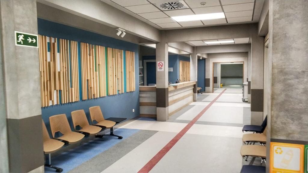 Un largo pasillo en un hospital con sillas y una señal de salida verde.