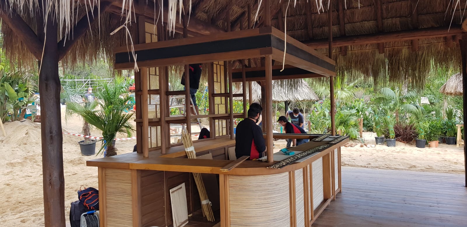 Un hombre está de pie detrás de una barra de madera bajo un techo de paja.