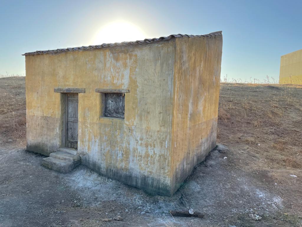 Una pequeña casa amarilla está sentada en el medio de un campo de tierra.