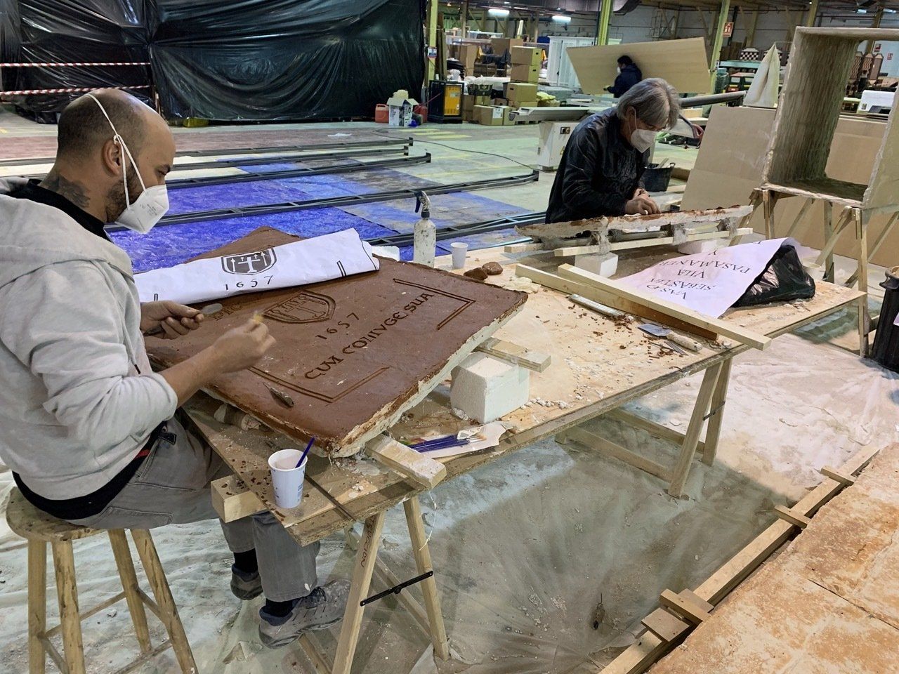 Dos hombres con máscaras trabajan en un trozo de madera en una fábrica.