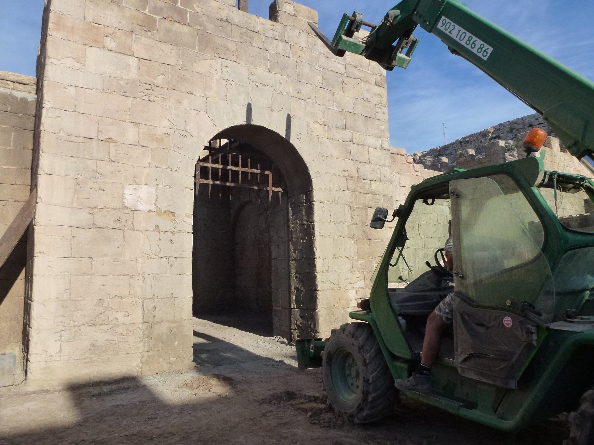 Un hombre conduce una carretilla elevadora verde frente a un muro de piedra.