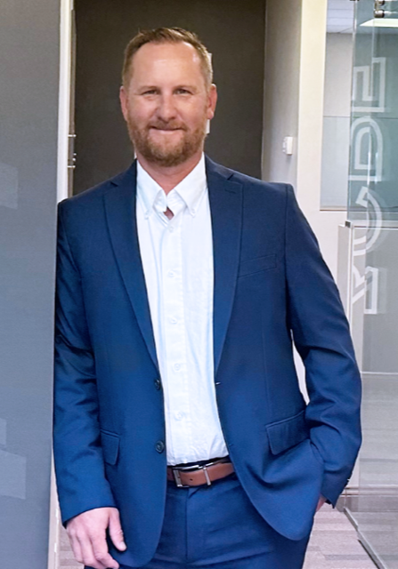 A man in a suit and blue shirt is standing in front of a tree.