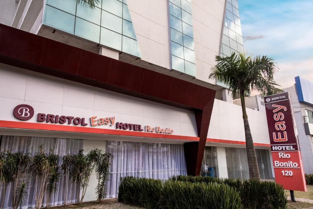 Hotel Bristol Easy no Brasil. Placa e detalhes vermelhos, prédio branco com fachada de vidro e palmeira na frente.
