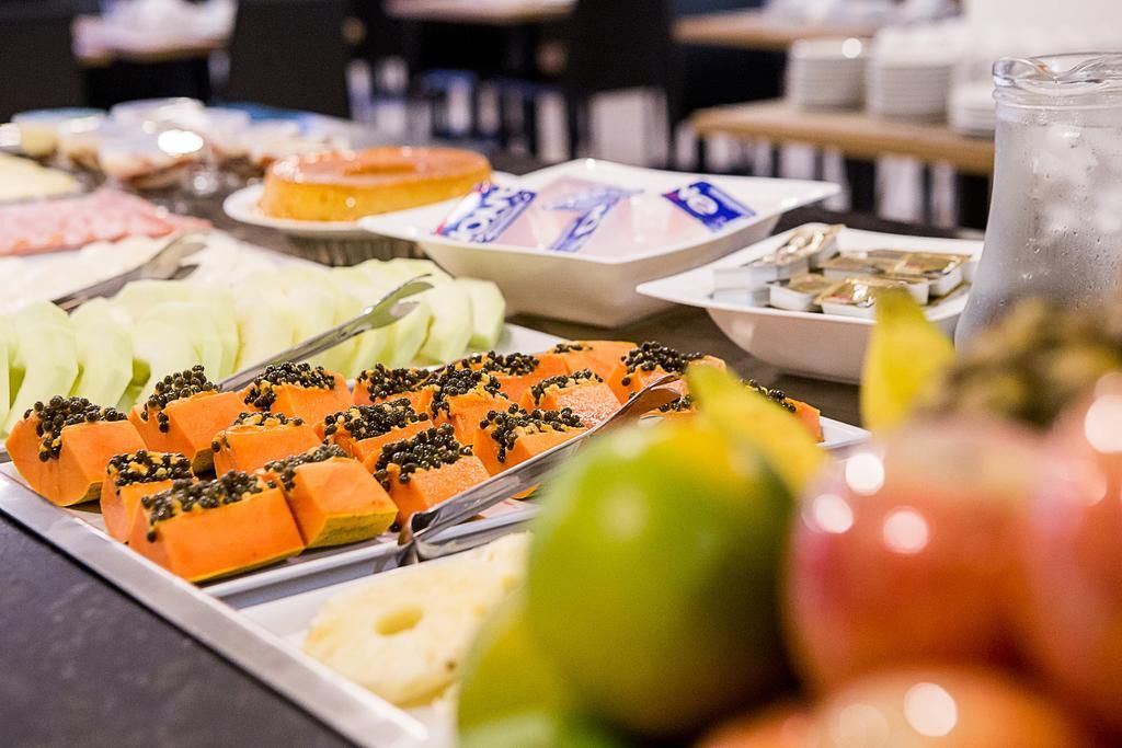 Buffet com frutas variadas, como mamão, maçã e abacaxi. Outros alimentos e uma jarra de água também estão disponíveis.