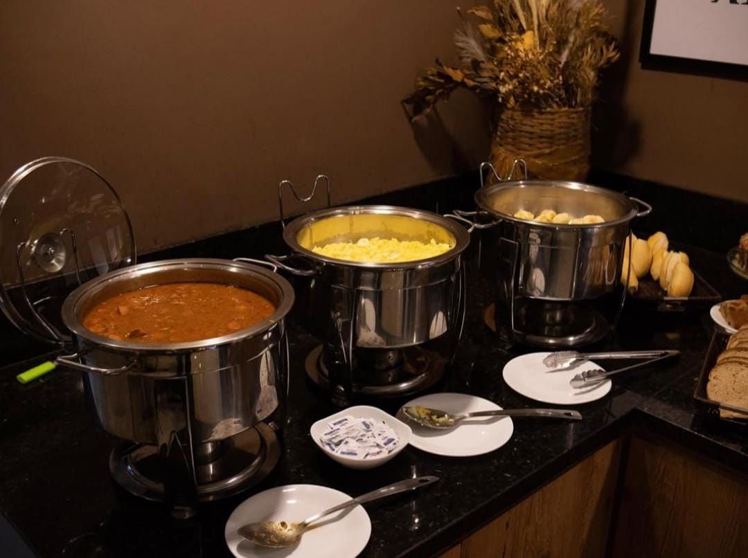 Um bufê com três pratos principais: chili, ovos mexidos e batatas, com utensílios para servir.