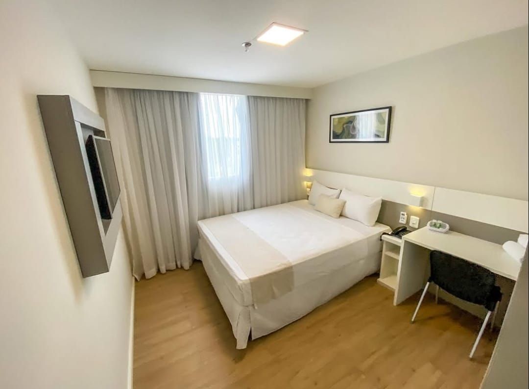 Quarto de hotel com cama, escrivaninha e janela com cortinas.