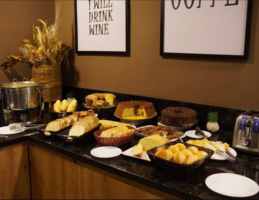 Buffet de café da manhã: Vários pães, bolos e doces em um balcão com cafeteira, placas e elementos decorativos.