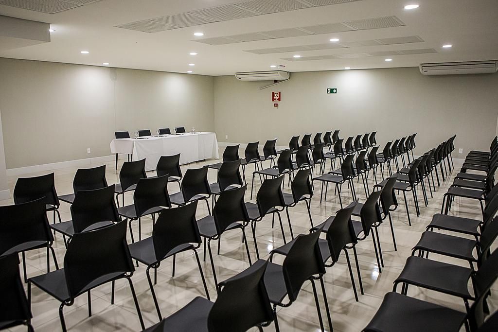 Fileiras de cadeiras pretas ficam de frente para uma mesa branca em um palco em uma sala de conferências.