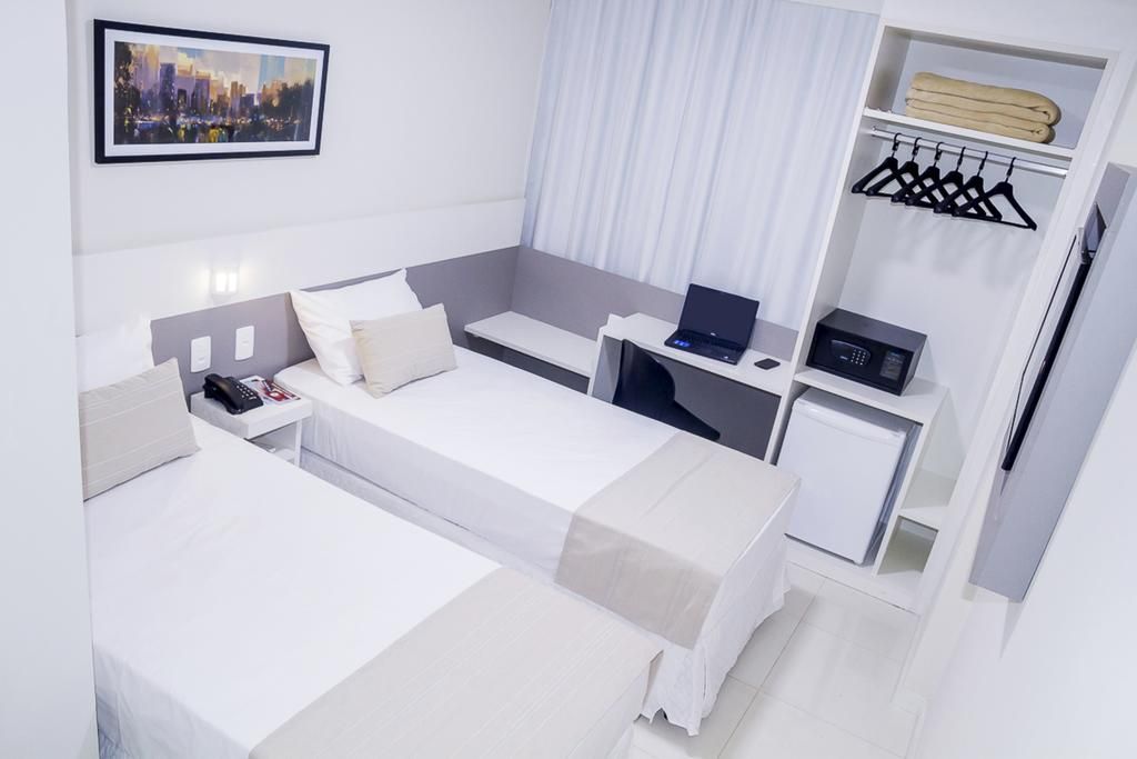 Quarto de hotel com duas camas, escrivaninha, guarda-roupa e obras de arte; predominantemente branco com detalhes em bege.