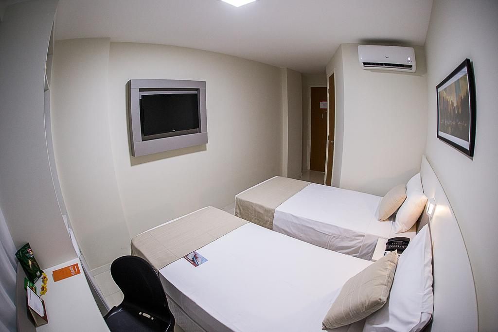 Duas camas em um quarto de hotel; TV na parede, quadros, ar condicionado e uma escrivaninha.