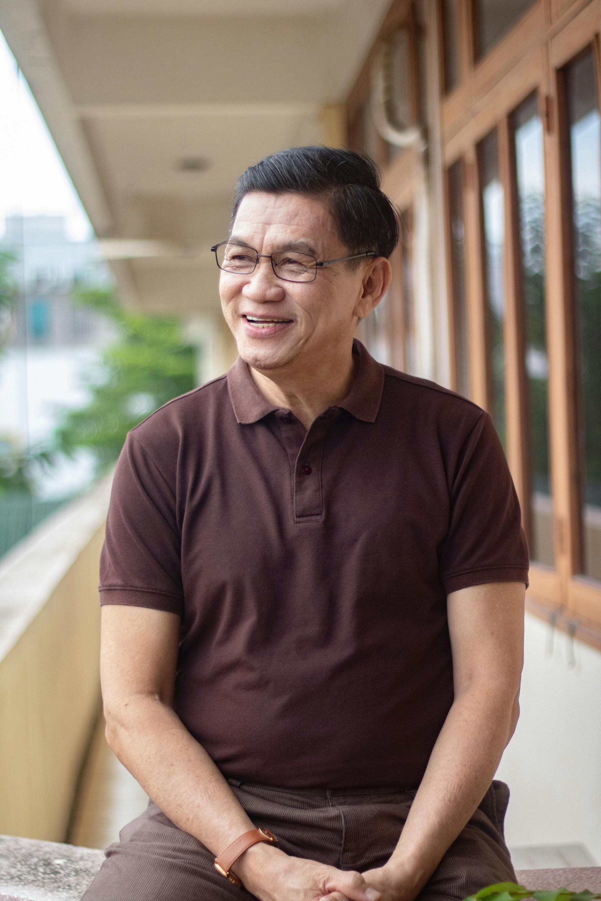 A man wearing glasses and a brown polo shirt is sitting on a balcony.