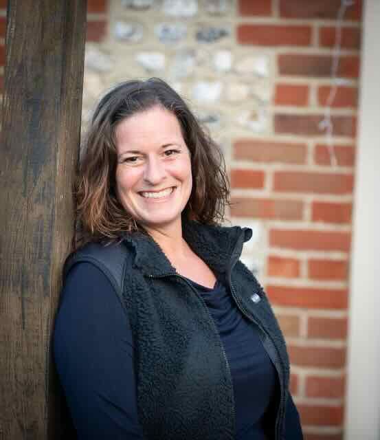 Photograph of Sarah Cutler leaning against a wooden post, smiling; a flintstone and brick wall background.