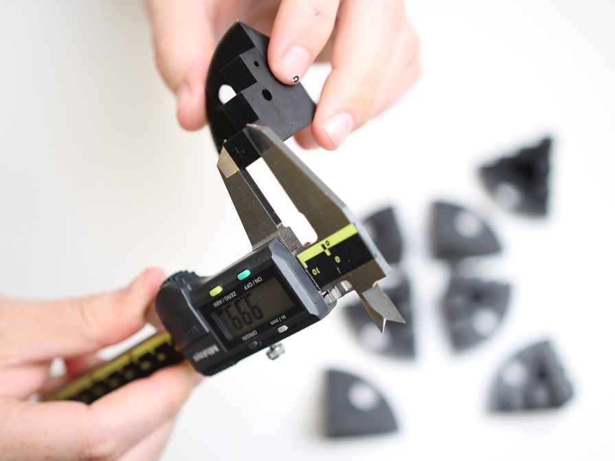 Hands using digital calipers to measure a black plastic piece, other pieces nearby.