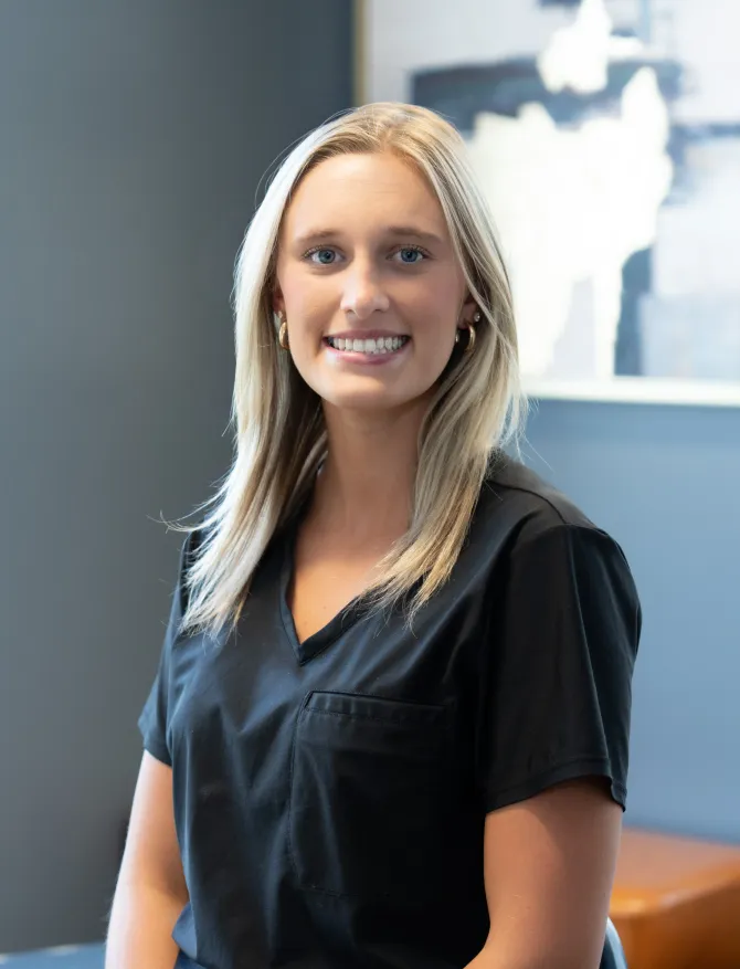 Blonde woman in black scrubs smiles at the camera in an office setting.