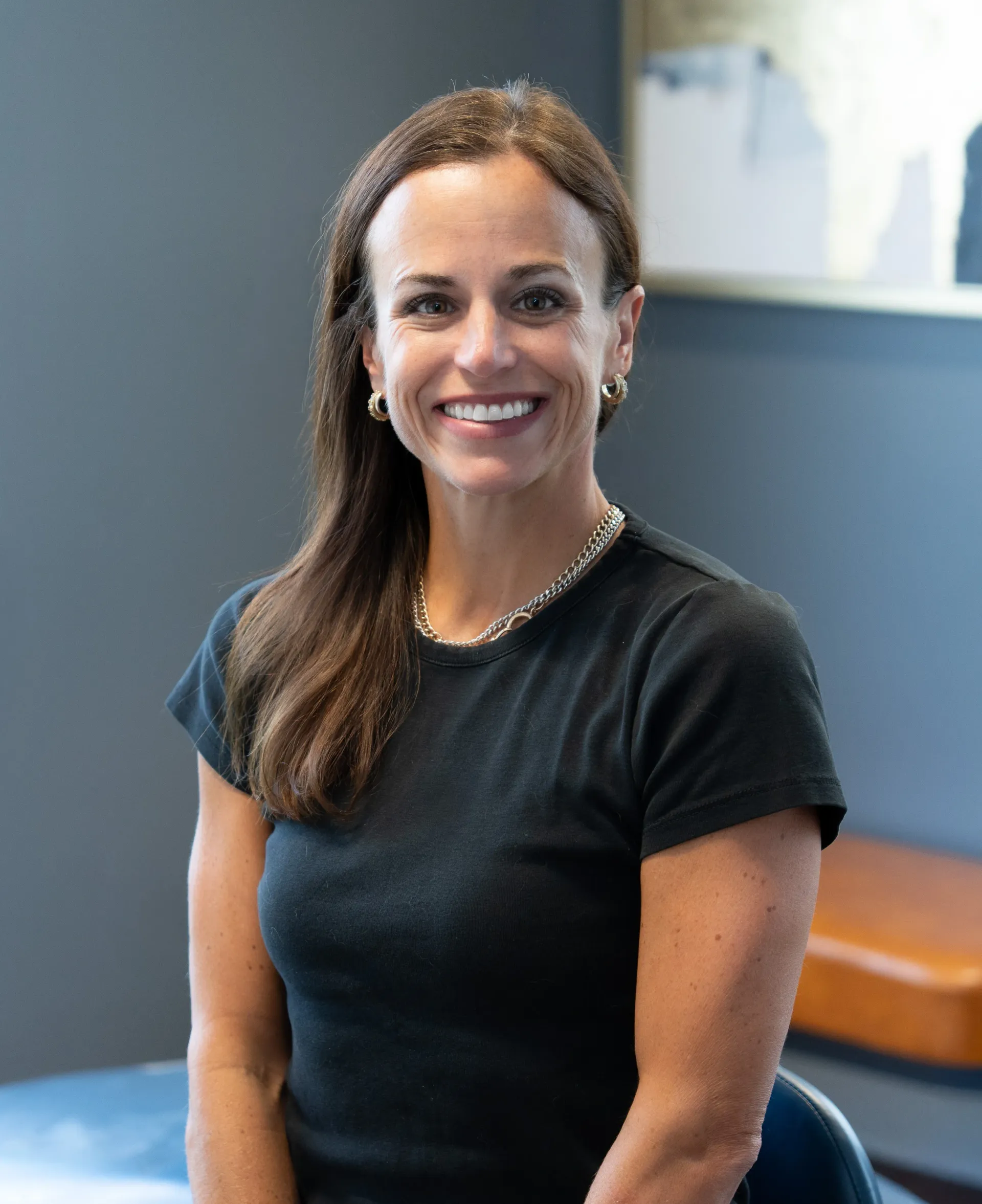 Woman with dark hair smiles, wearing black scrubs, dental office setting.