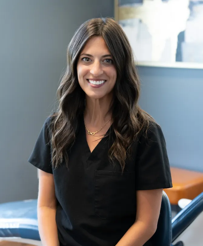 Woman smiling, wearing black scrubs, in a dental office setting.