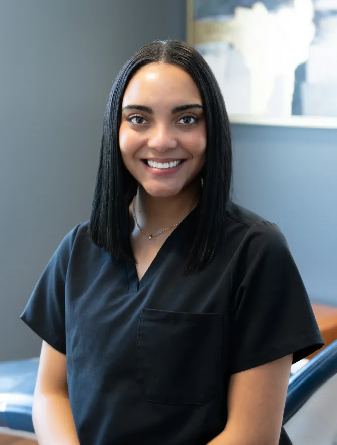 Woman with braided hair smiling in black scrubs; dental office setting.