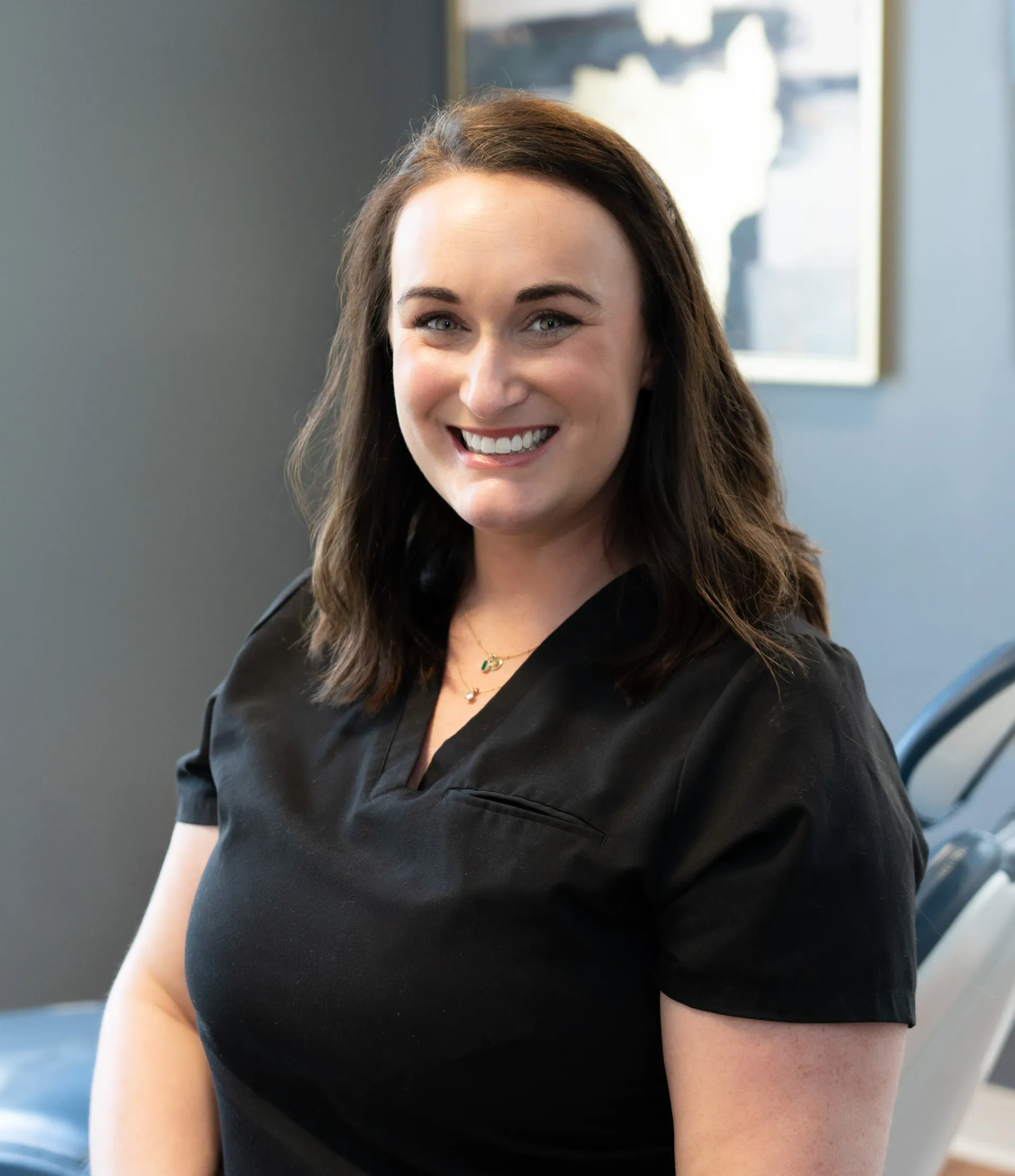 Woman smiles, wearing black shirt and silver necklace. Leans toward the camera, in a room with blue and orange accents.