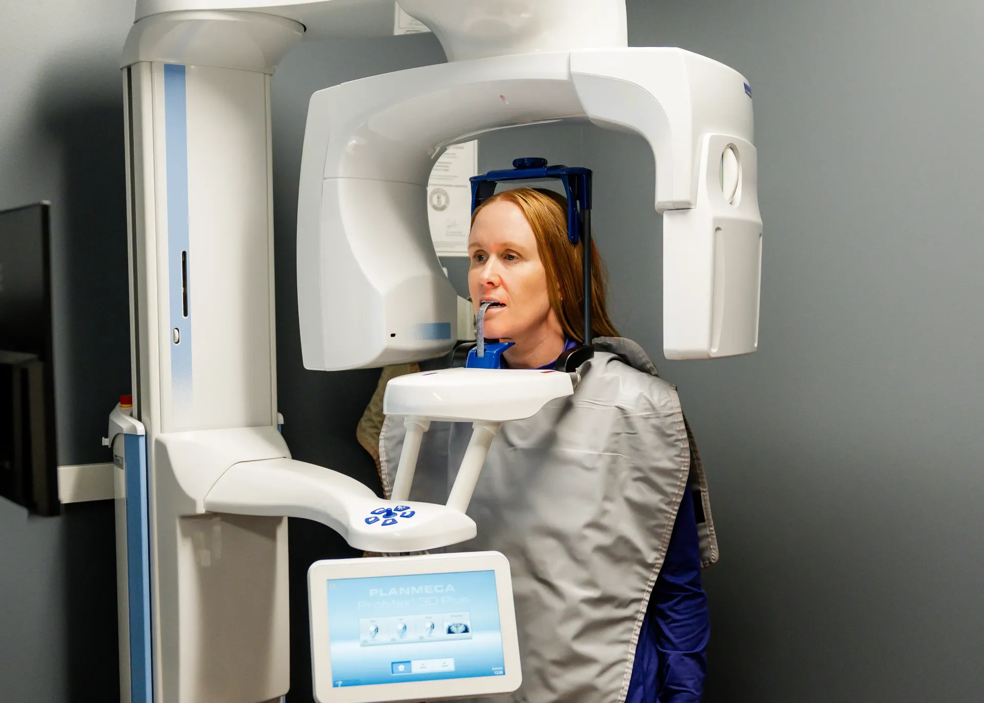 Woman in dental office having panoramic X-ray taken; white machine, gray apron.