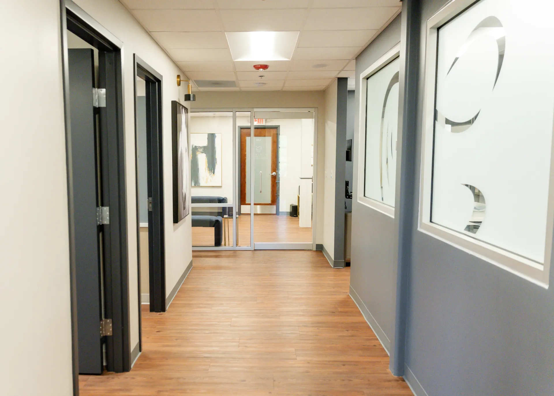 Hallway with doors, windows, and wood-look flooring. Light from ceiling fixtures illuminates the space.