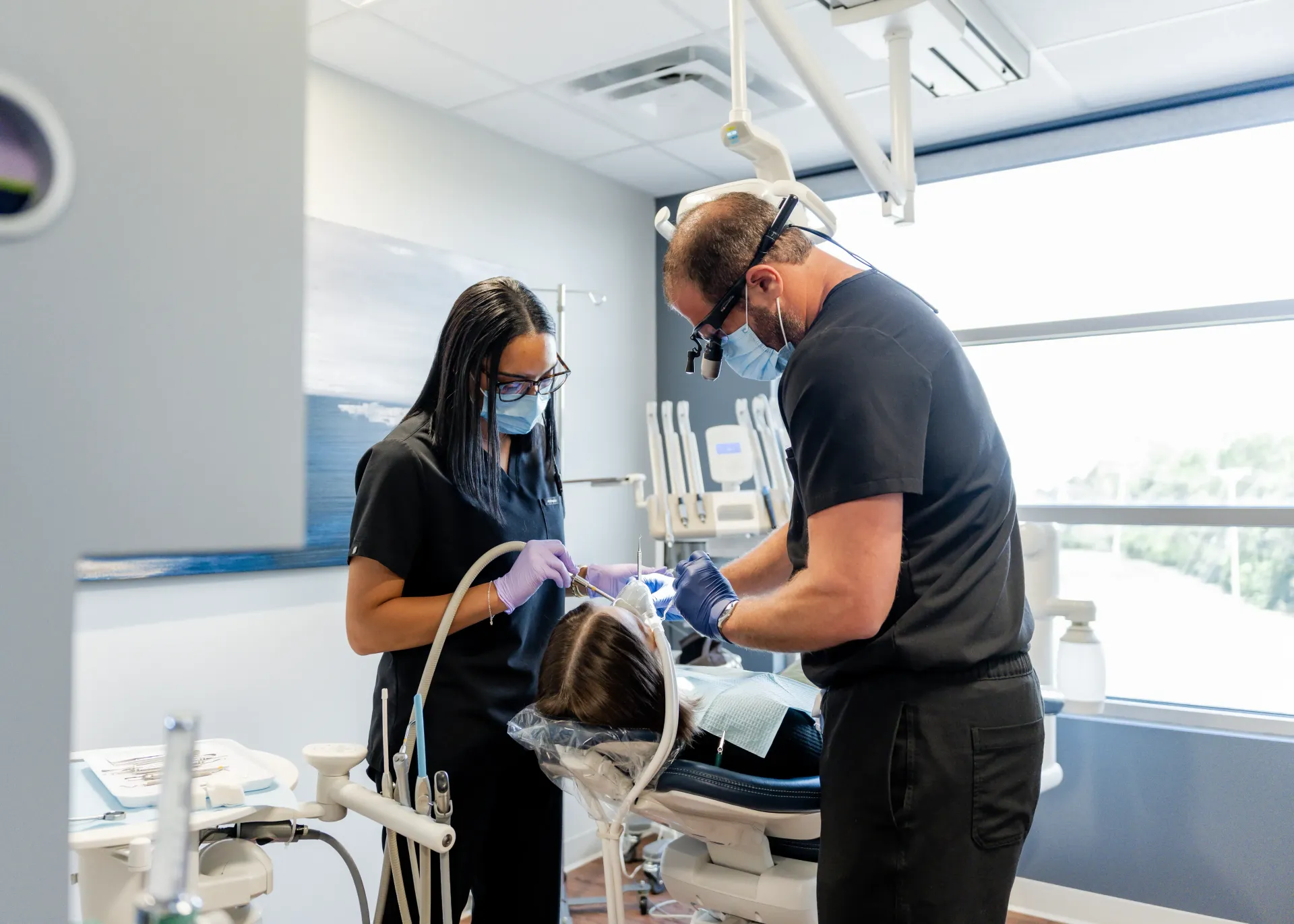 Two dental professionals wearing masks and scrubs examine a patient in a dental office.