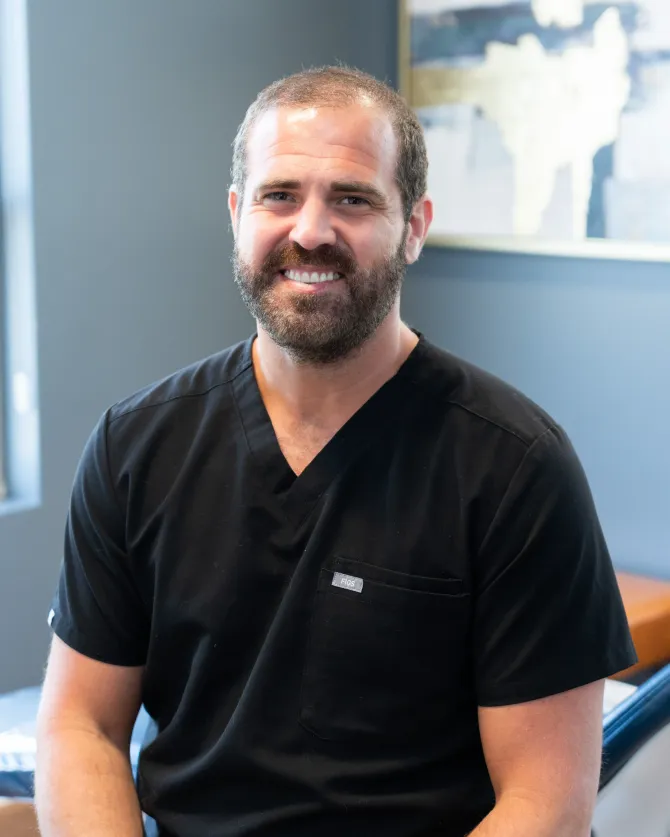 Man with a beard in black scrubs smiles at the camera, sitting inside a medical office.