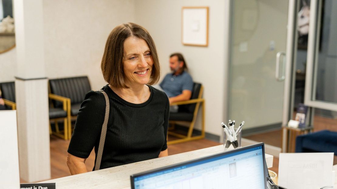 Woman smiles at a reception desk in a brightly lit office. A person sits in the background.