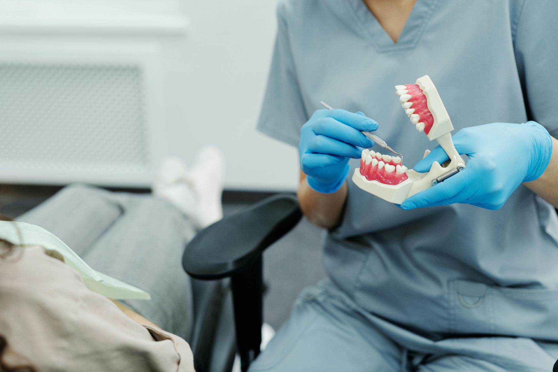 Dentist in blue scrubs demonstrating on a tooth model, patient in the chair.