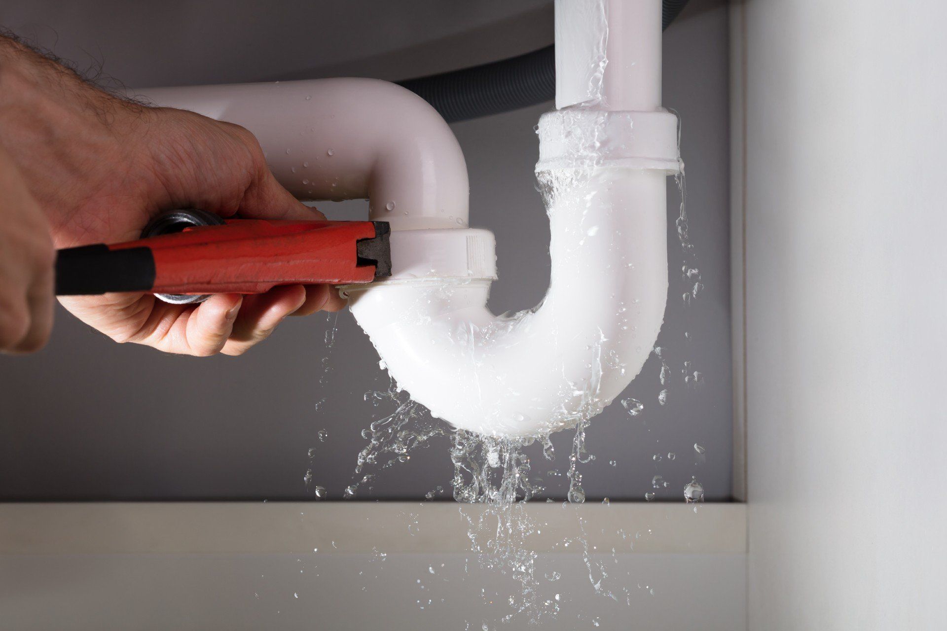 Hand tightening a wrench on a leaking white pipe under a cabinet.