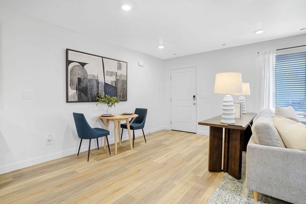 Open-plan living area with a small dining table, blue chairs, a beige sofa, and a window.
