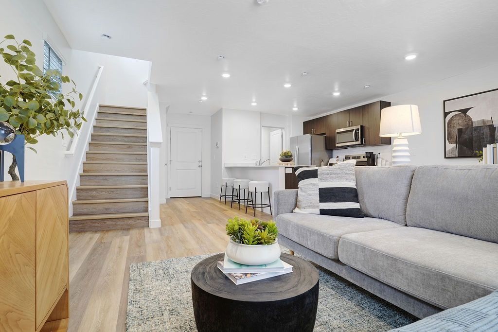 Open-concept living area with gray sofa, stairs left, kitchen island, and bright white walls.