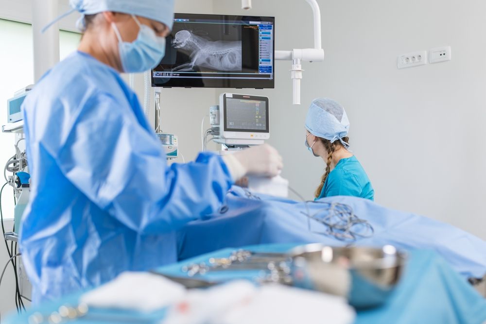 Surgeons in blue scrubs operating in a sterile surgical room, with equipment and a screen displaying an x-ray.