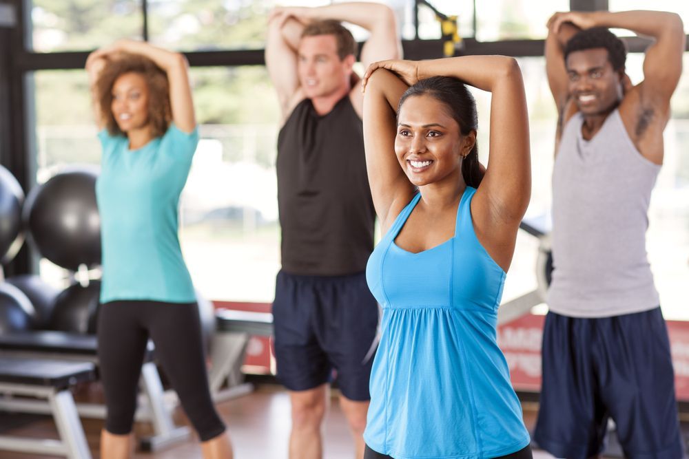 Group of people smiling, stretching arms overhead in a gym, wearing workout clothes.