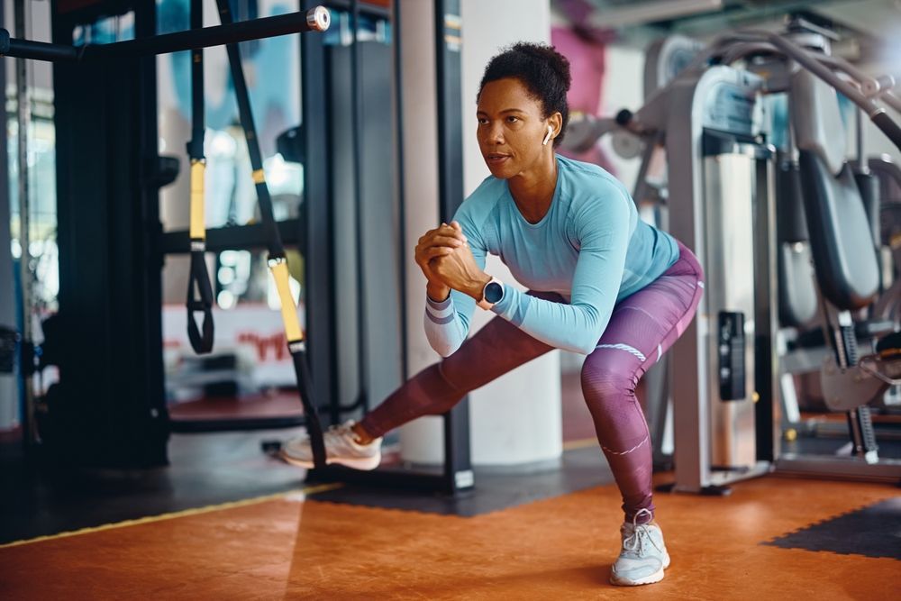 Woman doing a side lunge in a gym, wearing blue shirt and purple leggings.