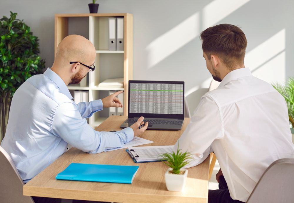 Two men are sitting at a table looking at a laptop computer.