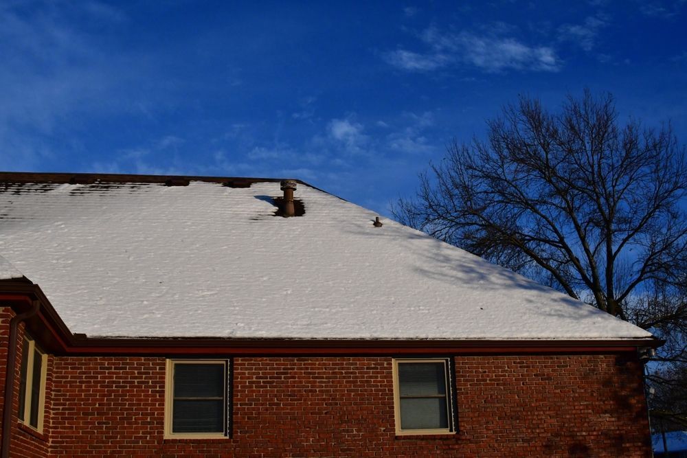 A brick house with snow on the roof and a tree in the background.