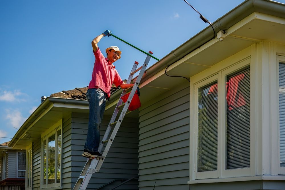 A man is standing on a ladder cleaning the gutters of a house.