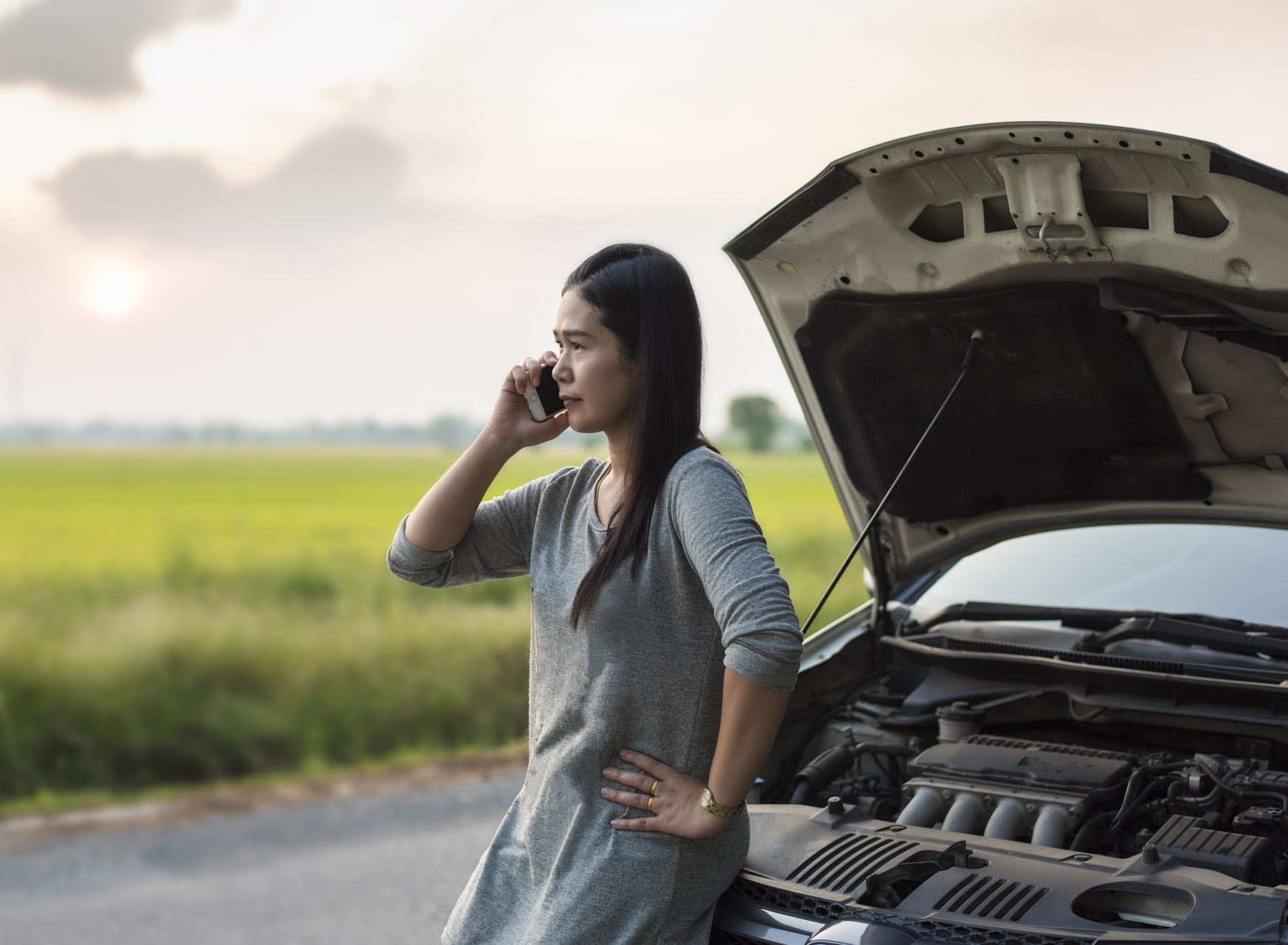 the owner of the car stands on the side of the road with her front hood open, she is on the phone with her insurance company discussing what to do with her totaled car that is still drivable