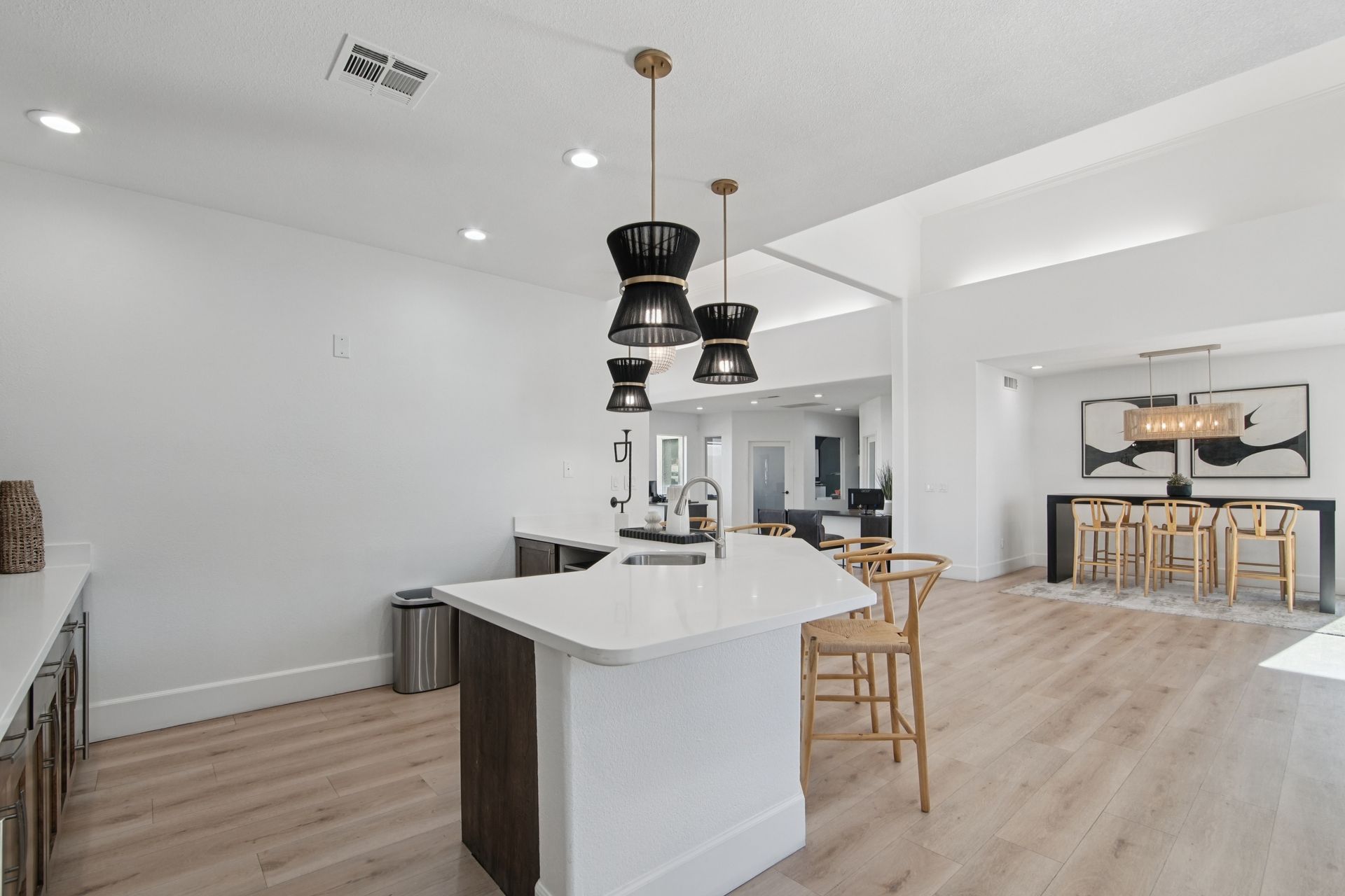 Modern kitchen with island, pendant lights, and light wood floors. White walls and dining area in the background.