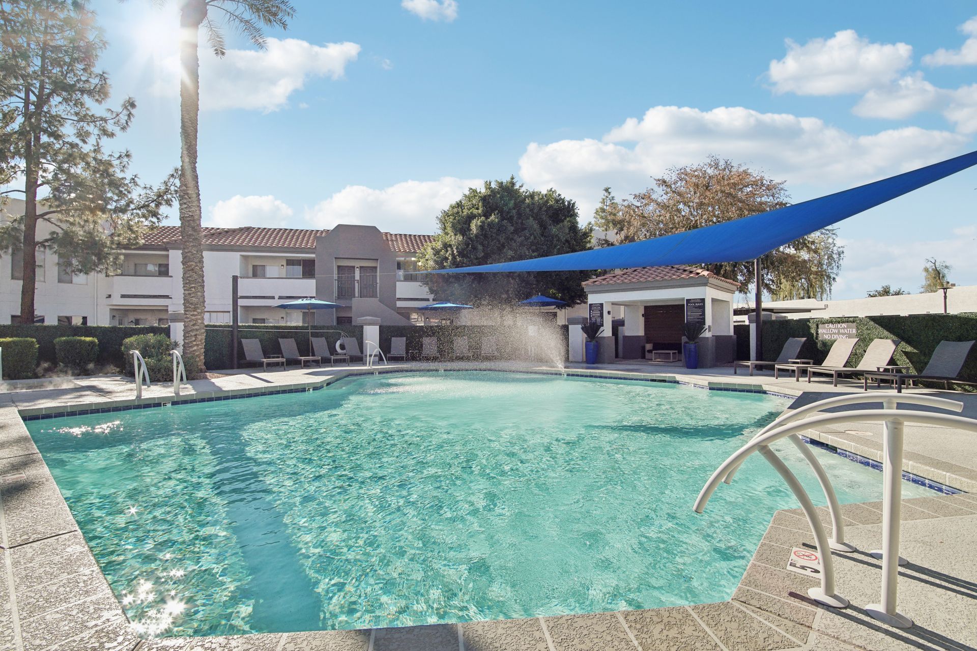 Swimming pool with fountain and shaded lounge area under blue canopy.