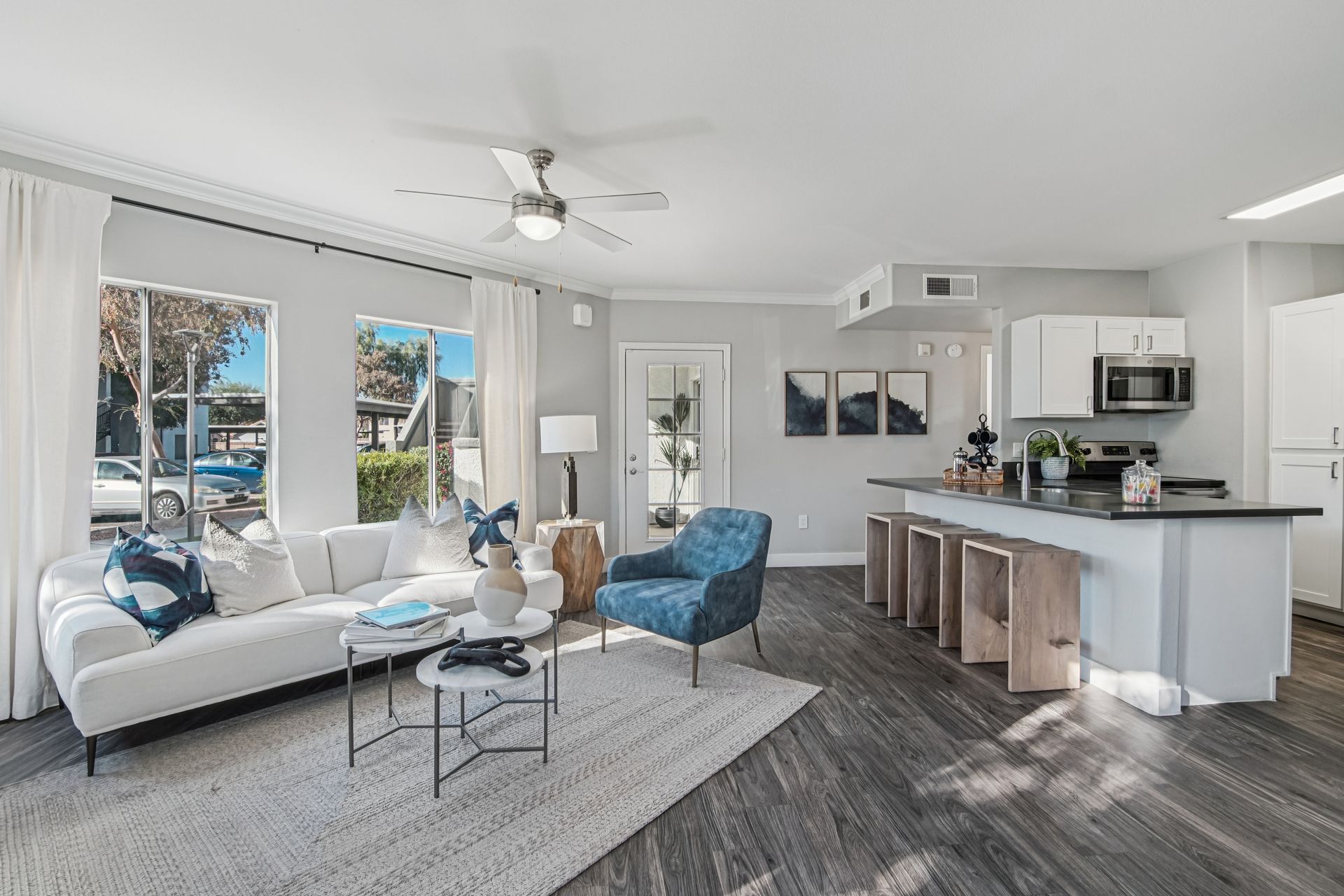 Living room with white sofa, blue chair, open kitchen with bar seating, and large windows.