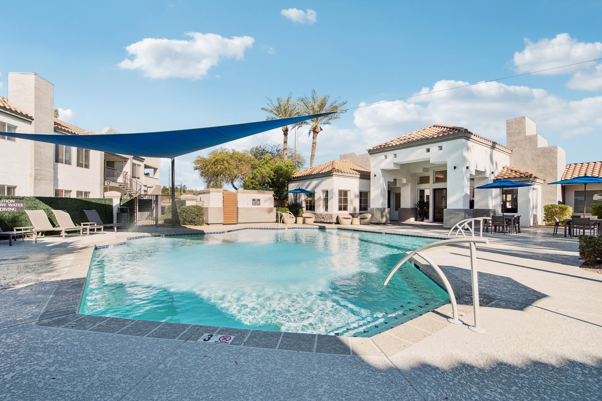 Swimming pool area with blue water, lounge chairs, and buildings under a sunny sky.