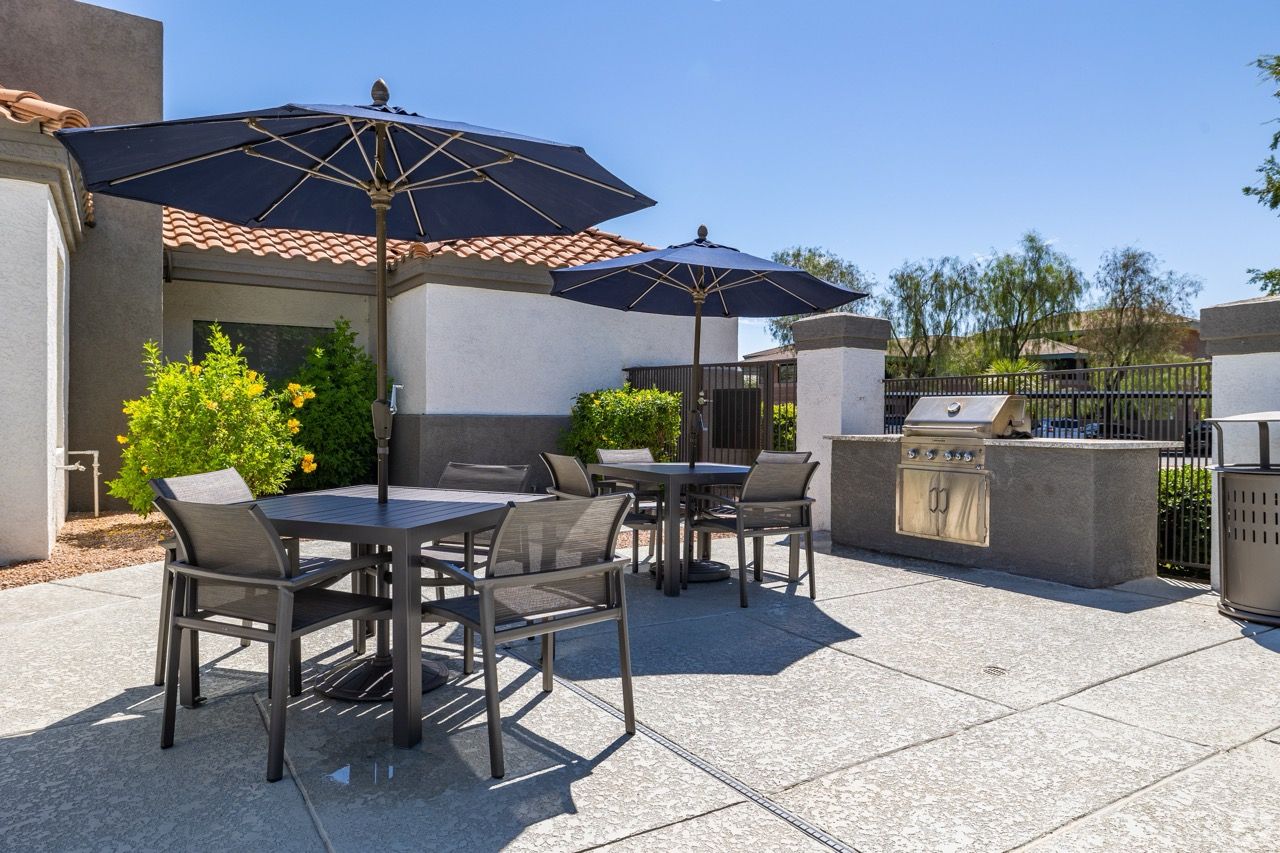Outdoor communal patio with tables, chairs, and large umbrellas beside a built-in grill.