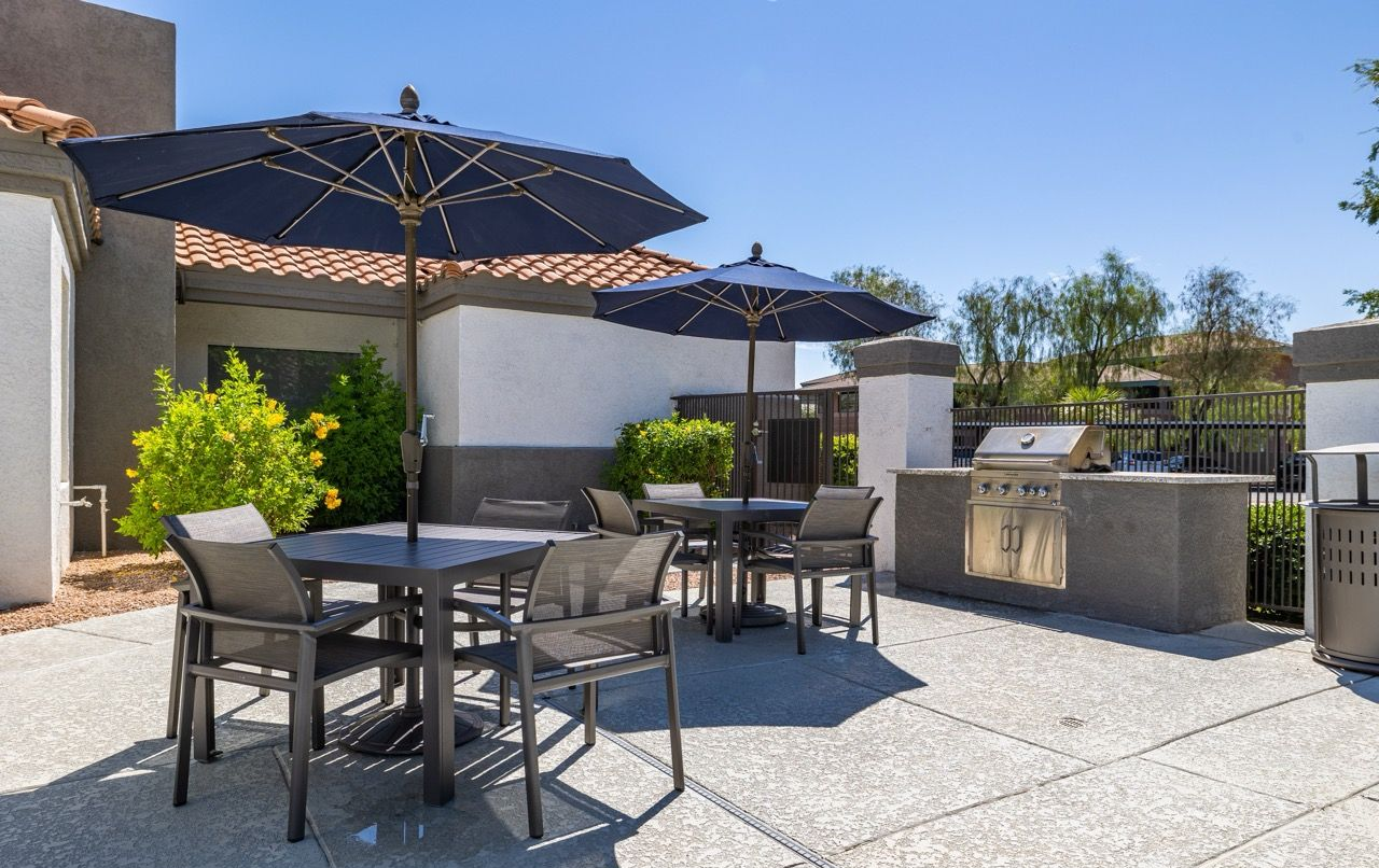 Outdoor communal patio with tables, chairs, and large umbrellas beside a built-in grill.