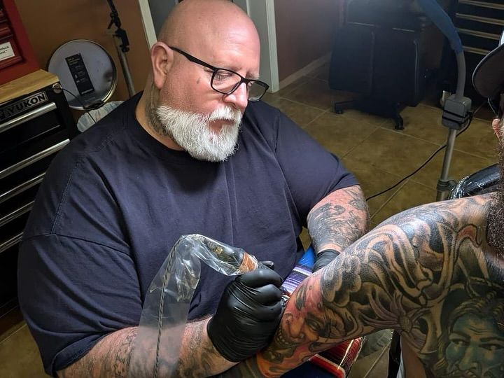 Tattoo artist with a gray beard and glasses tattoos a person's arm, indoors.