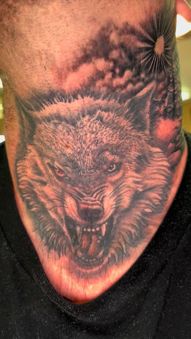 Neck tattoo of a snarling wolf's face under a sun and cloudy sky; the tattoo is on a person's neck.