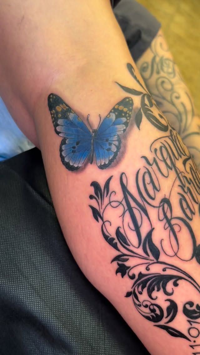 Blue butterfly tattoo on an arm, above ornate black script and flourishes.