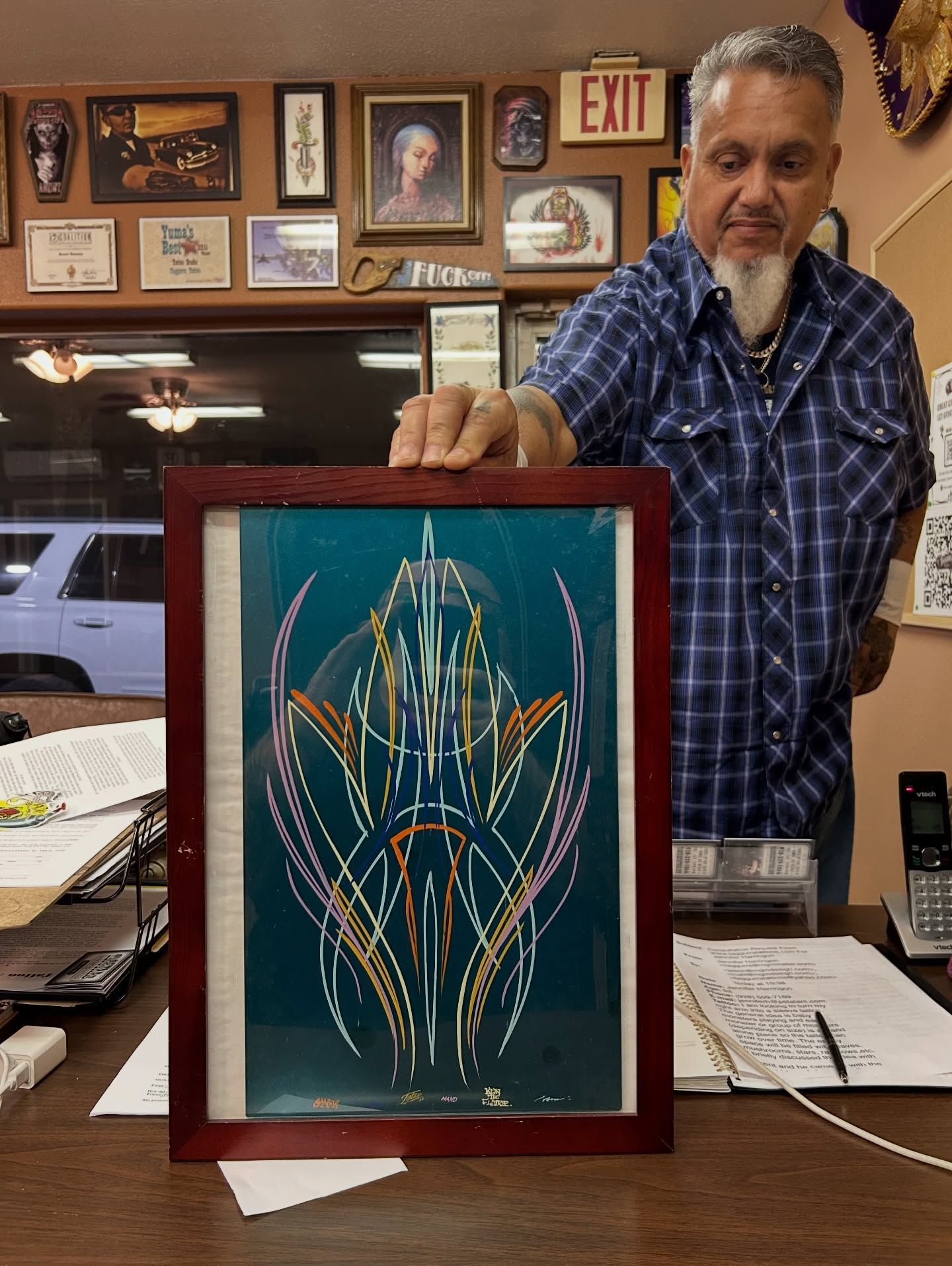 Man holding framed pinstripe art. The art has teal background with gold, pink, and white lines. Inside a shop.