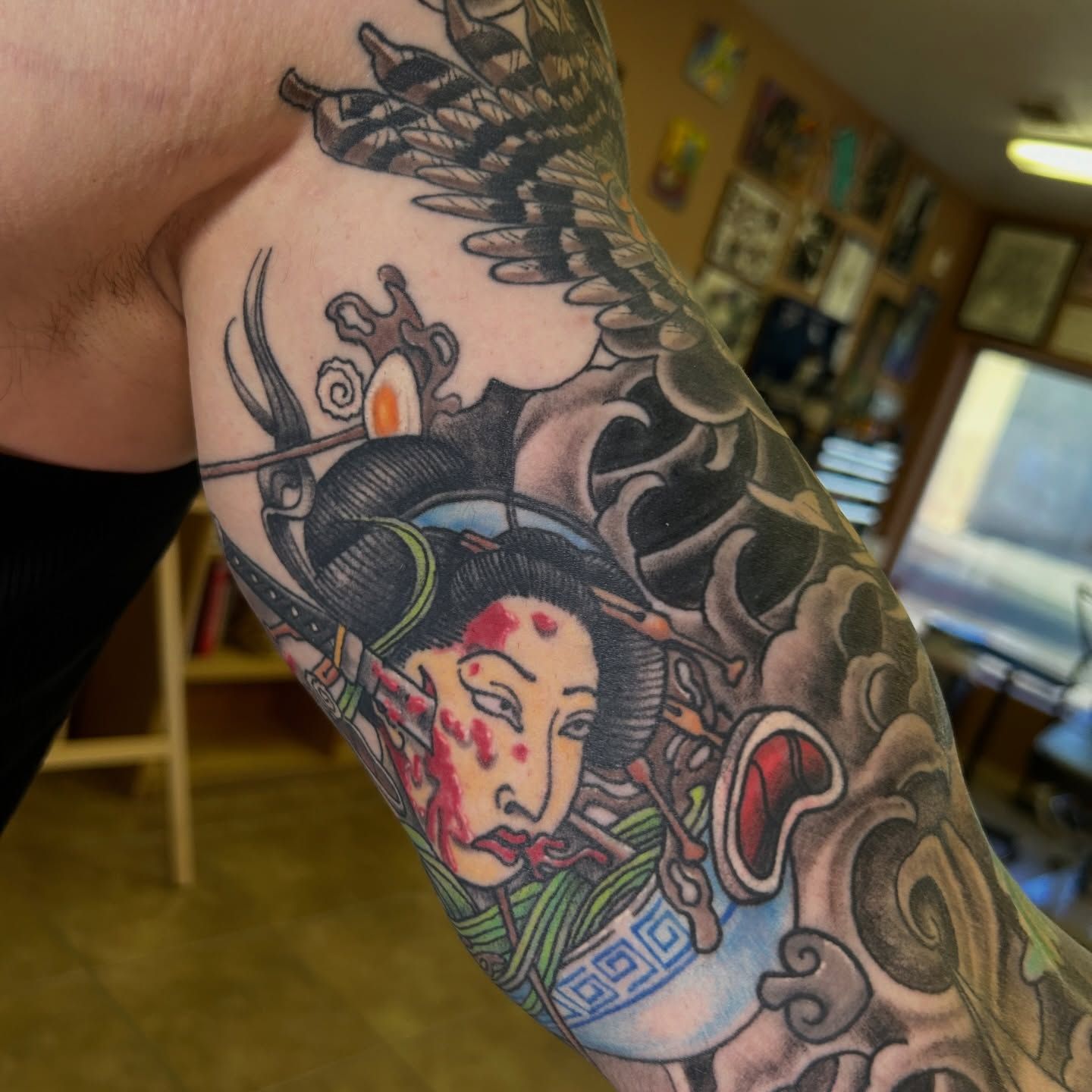 Tattoo of a geisha with red blood on face, in a sleeve design. Part of an eagle and cloudy background.
